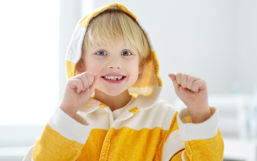 Smiling blonde toddler in yellow and white hoodie with hands up in bright indoor setting.